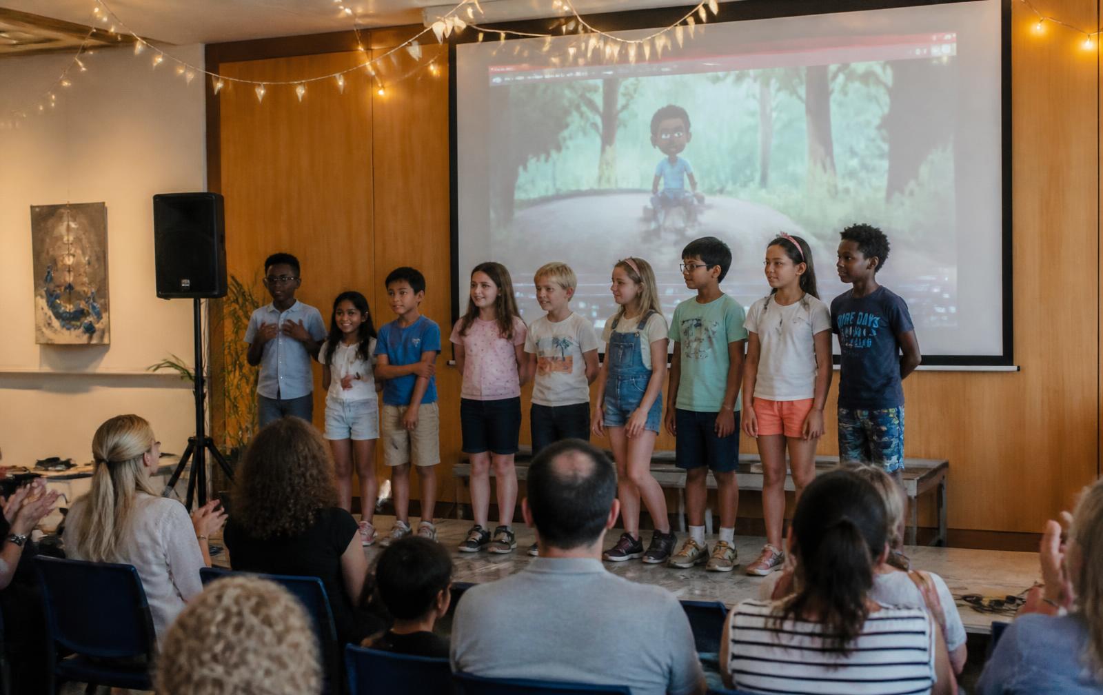 Children on stage presenting their animated movie on a big screen while parents applaud at the end-of-camp showcase